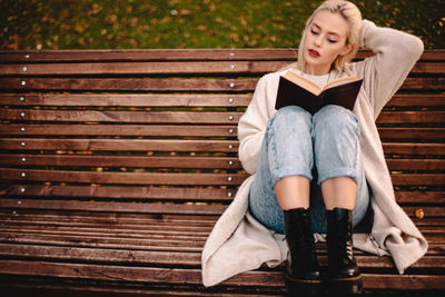 Young woman reading book sitting on bench in park during autumn