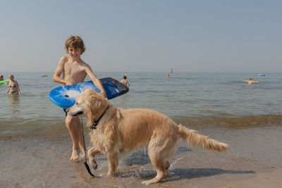 Shirtless boy carrying surfboard walking with golden retriever walking at beach against sky