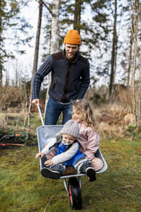 Playful man wheeling son and daughter sitting in wheelbarrow at back yard