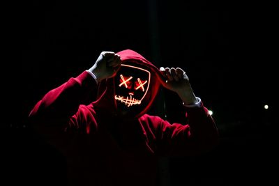 A man wearing a red hoodie with red glowing mask in the dark backgroun