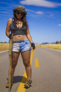 Portrait of young woman in shorts standing with stick on country road against sky