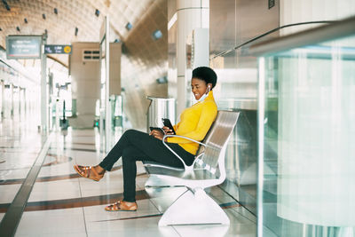 A young african woman in a protective mask sits in a chair in the waiting room, listening to music 