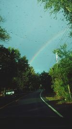 Empty road amidst trees against rainbow in sky