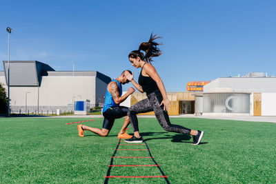 Full body side view of focused sporty man and woman doing lunges while exercising with agility ladder on green grassy lawn on sports ground