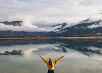 Rear view of unrecognizable woman with arms outstretched up looking at mountains reflected in lake