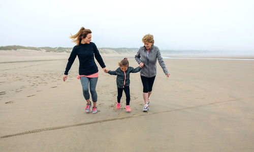 Playful woman walking with mother and daughter at beach against sky