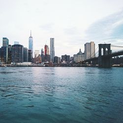 City skyline and brooklyn bridge by river against sky