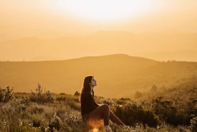 Side view of young woman sitting on land against sky during sunset