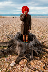 Unrecognizable person in red hat and coat standing in stump center on shore with crushed stones in summer