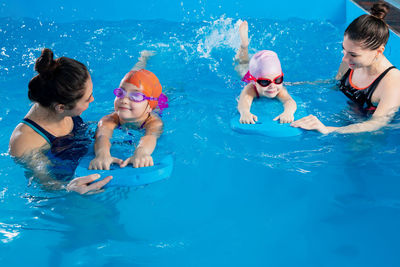 Two trainers teaching little girls how to swim in indoor pool with pool floating board