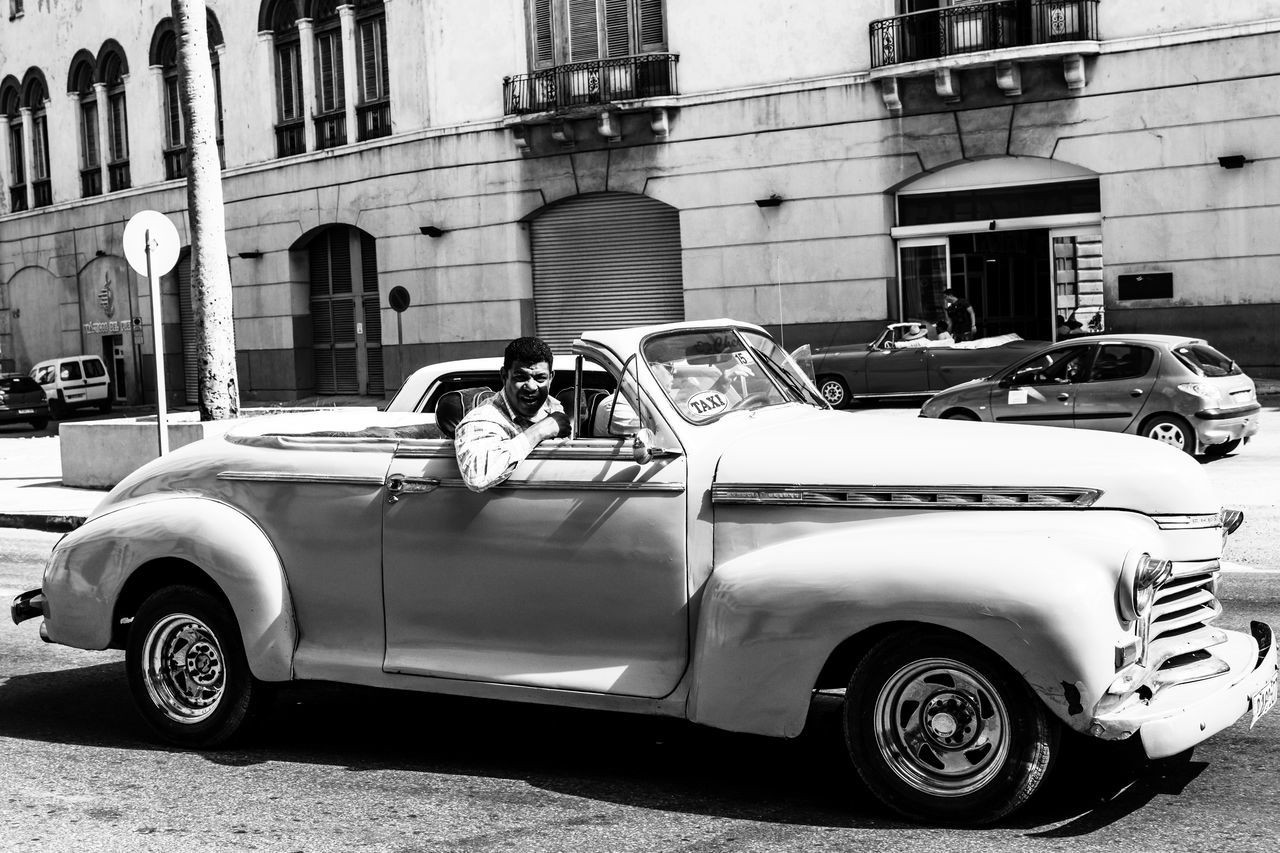 car, vehicle, mode of transportation, land vehicle, transportation, motor vehicle, architecture, building exterior, built structure, city, automobile, street, retro styled, vintage car, day, antique car, luxury vehicle, black and white, mid-size car, the past, history, building, travel, old, road, automotive exterior, outdoors, monochrome photography