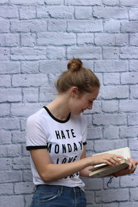 Smiling teenage girl reading book while standing against brick wall