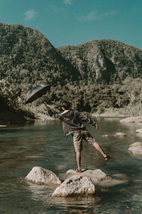 Rear view of man holding umbrella amidst river on rock