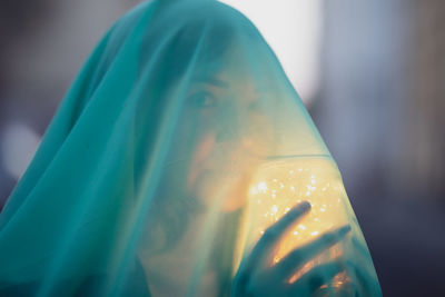Close-up portrait of woman holding illuminated jar under blue scarf at dusk