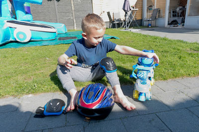 Blonde caucasian child puts on blue roller skates,sits on the green lawn in the courtyard