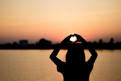 Silhouette woman making heart shape with hands by lake against sky during sunset