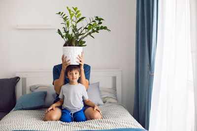 Bearded father and son in t-shirts are sitting on a bed. man is holding pot with flower on his head