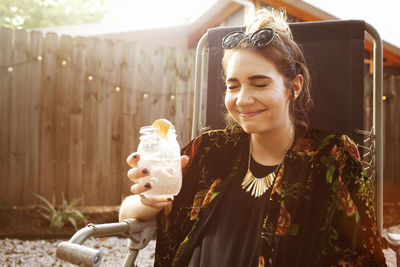 Happy woman with eyes closed holding drink jar while sitting in yard