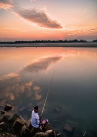 Man fishing in lake against sky during sunset