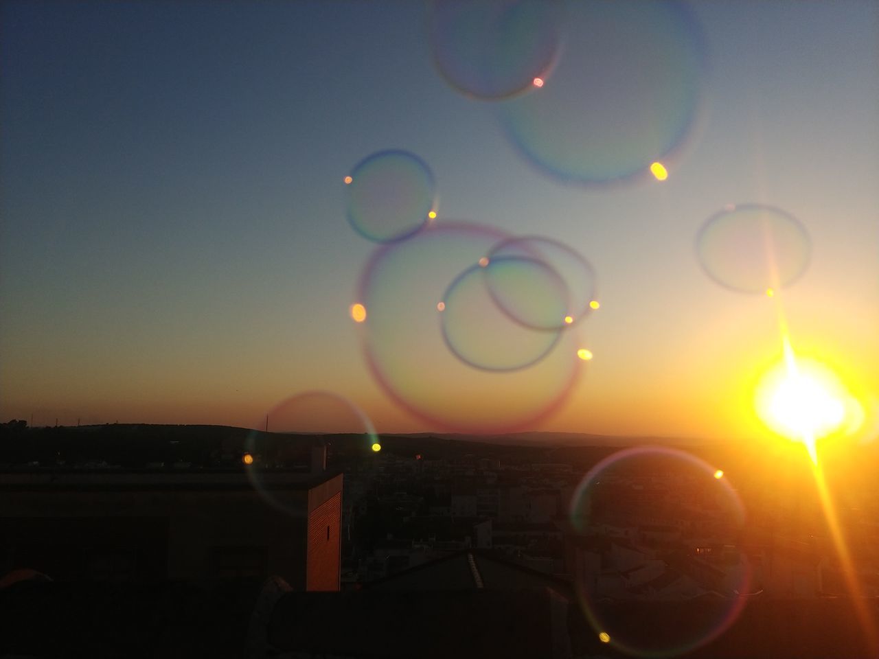 sky, light, bubble, mid-air, nature, sunset, flying, sunlight, no people, cloud, reflection, lens flare, astronomical object, liquid bubble, outdoors, lighting, multi colored, fragility, yellow, soap sud, sun, beauty in nature, transparent, bubble wand, shape, circle, tranquility, balloon, evening, geometric shape, clear sky, environment, architecture, sphere, orange color