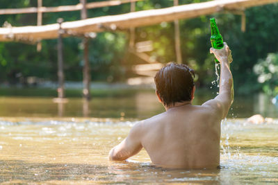 Rear view of shirtless man holding bottle in river