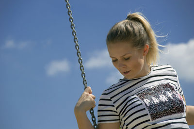 Low angle view of young woman looking down while swinging against sky