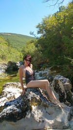 Young woman sitting on rock by tree against sky