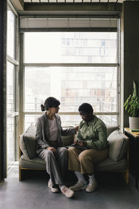 Full length of mature businesswoman consoling female colleague sitting on sofa against window at office