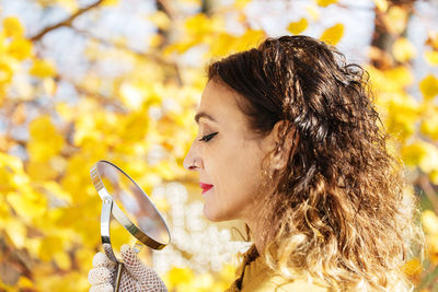 Close-up of mature woman holding hand mirror standing by autumn tree