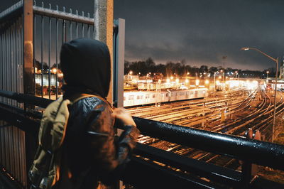 Rear view of person standing by railing over illuminated railroad track against sky at night