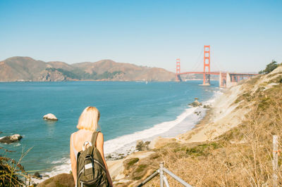 Rear view of woman standing at sea shore by golden gate bridge against clear blue sky