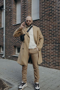 Portrait of man talking on phone while standing against building on footpath