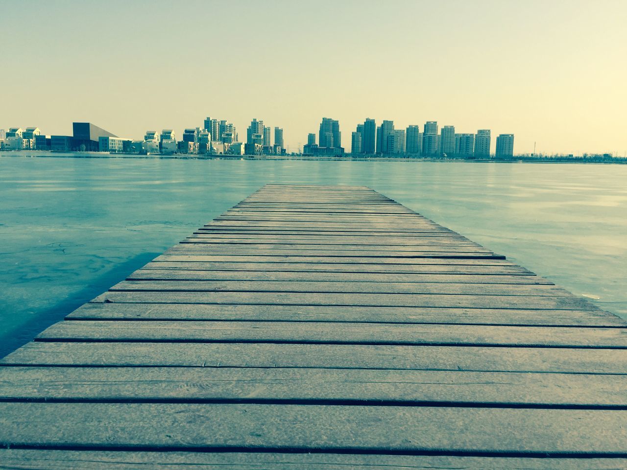 clear sky, built structure, architecture, building exterior, copy space, the way forward, city, surface level, pier, diminishing perspective, wood - material, water, skyscraper, in a row, outdoors, no people, day, tall - high, sea, sunlight