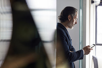 Businessman using smart phone while leaning on glass wall in office
