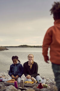 Smiling mother and daughter cooking on barbecue grill at beach against sky