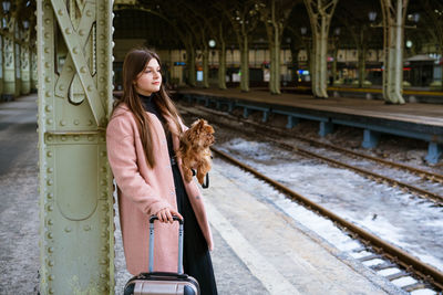 Travel concept. at station, young tourist with dog goes and drags suitcase