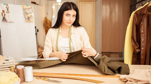 Black haired seamstress in atelier, portrait at work. table with fabric and sewing tools, indoors.