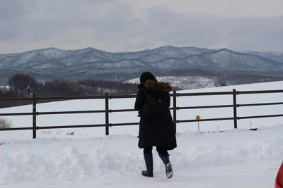 Rear view of woman walking on snow covered field against cloudy sky