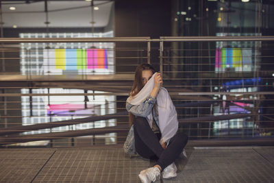 Woman holding fabric over face while sitting on footbridge against railing