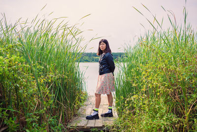 A brunette girl in a dress and a black jacket stands on the river bank on a wooden bridge