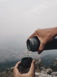 Cropped hands pouring drink in lid against landscape during foggy weather