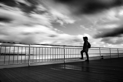 Full length of silhouette man standing on footpath while looking at sea against cloudy sky