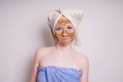 Portrait of young woman with facial mask wrapped in towel against white background