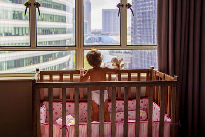 Rear view of girl standing on crib by window