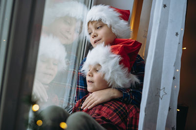 Children wearing santa hats and plaid shirts sitting hugging by the window waiting for christmas