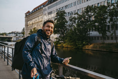 Happy male food delivery person standing near railing by canal in city