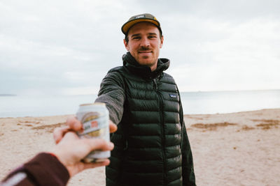 Cropped hand giving drink can to man standing at beach against sky