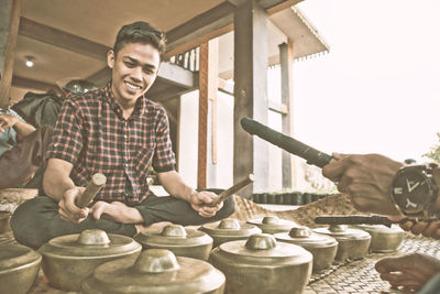 Smiling young man playing singing bowls while sitting against house