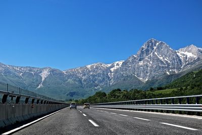 Cars on road leading towards mountains against clear blue sky during sunny day