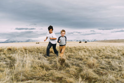 Happy brothers running while playing on grassy field against cloudy sky